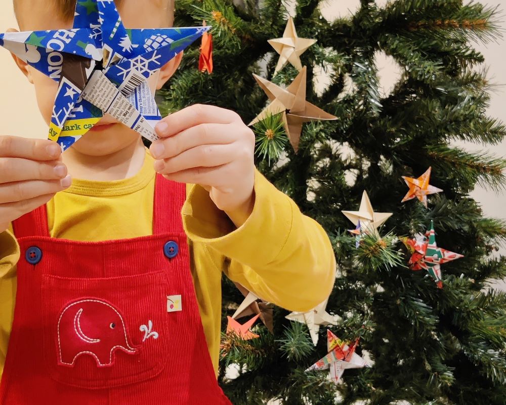 Child holding a blue origami star in front of a decorated Christmas tree. 