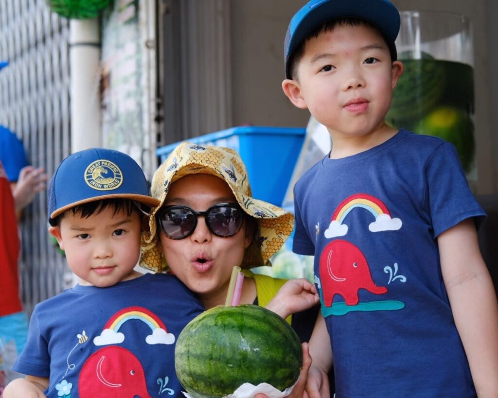 Liz is wearing sunglasses and a DUNS sunhat holding a water melon with her two boys either side in blue Babipur t-shirts