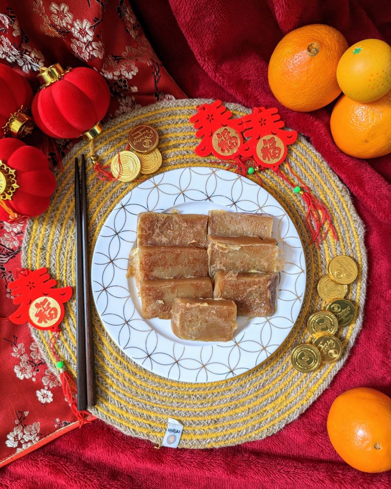 Slices of New Year's Cake on a decorative white and gold plate, surrounded by gold chocolate coins, chopsticks and oranges for Lunar New Years celebrations.