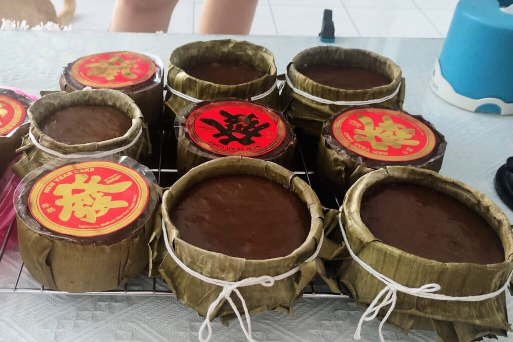 A selection of New Years cakes for Lunar New Year on a wire rack within banana leaves, some with decorative red lids with gold lettering.