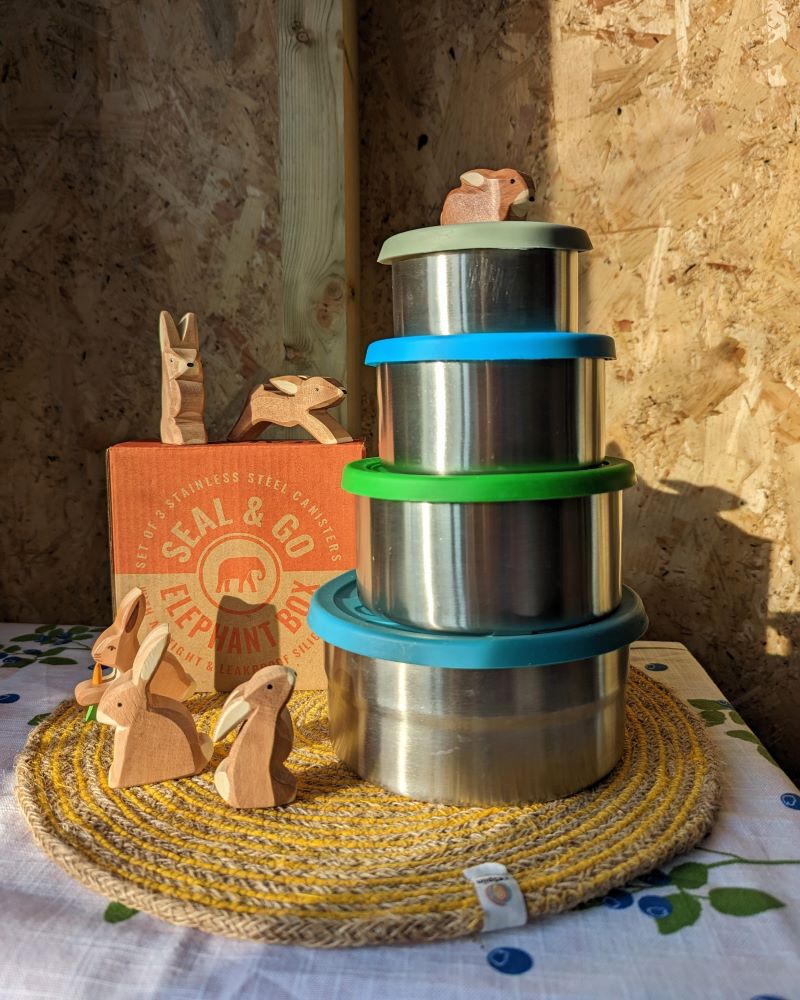 A stack of four circular metal containers with silicon lids on a woven placemat with wooden rabbit figures surrounding the tins.