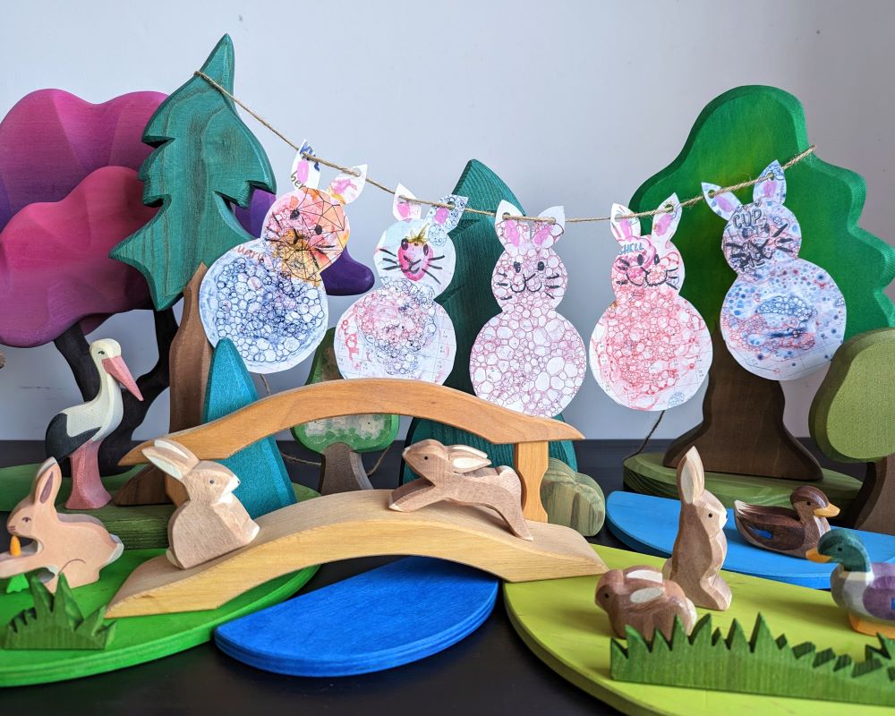 Liz has created a Lunar New Year, Year of the Rabbit play scene using wooden rabbit figures with a bridge over a stream and wooden trees in the background, with DIY rabbit bunting behind. 