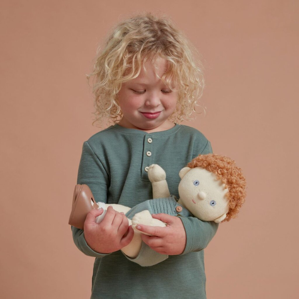 A child with curly blond hair holding Dinkum Doll Pea who has curly red hair and is wearing a blue knitted romper.