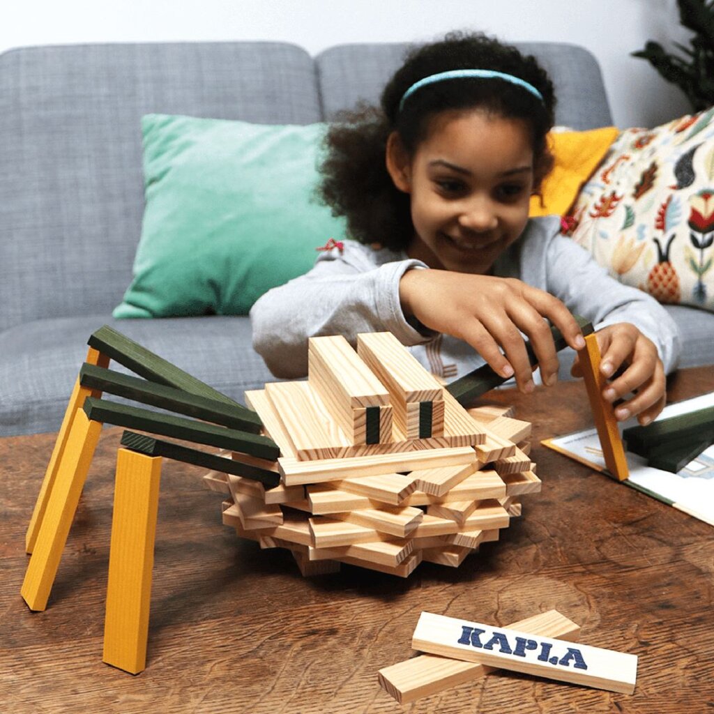 A child creates a Kapla Spider on a coffee table with a sofa behind