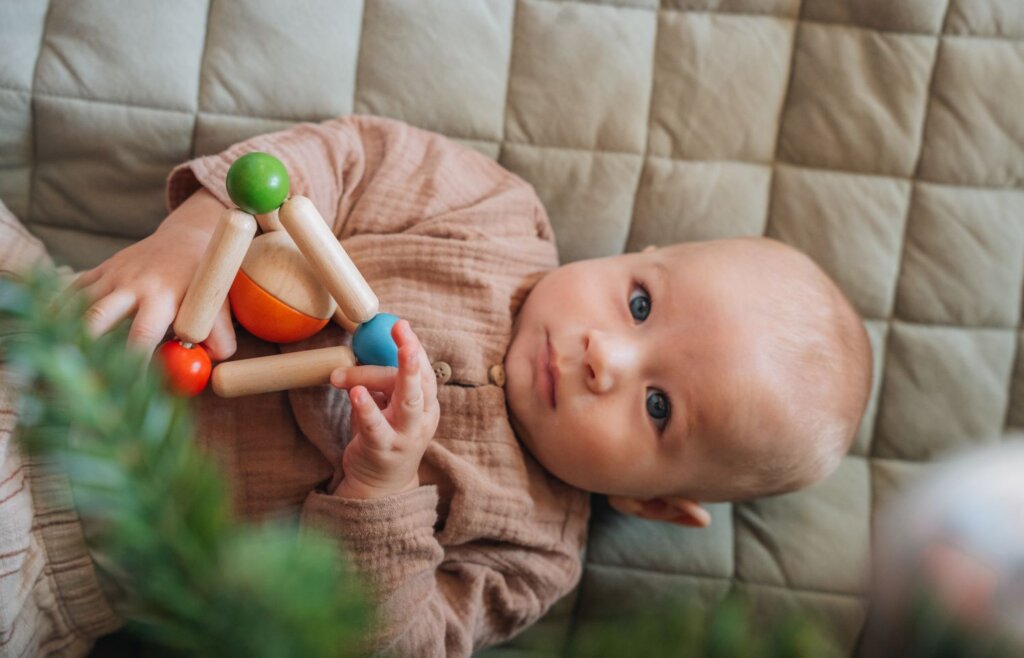 A baby on a blanket looking up through the Christmas Tree holding a wooden toy by PlanToys