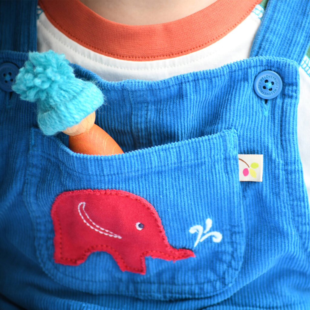 A peg doll wearing a woolly hat in the pocket of a child wearing blue Babipur dungarees